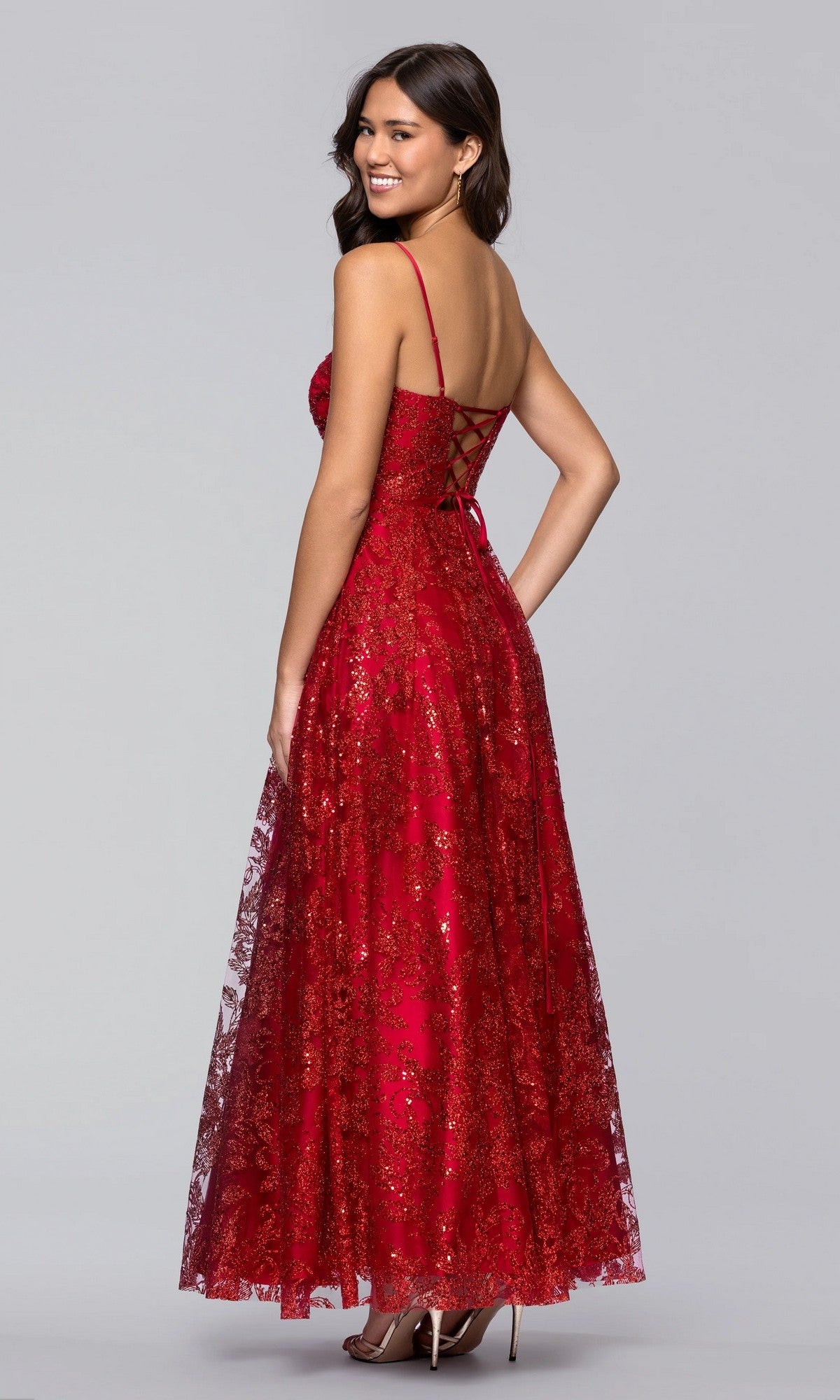 A woman poses and smiles against a gray background, wearing the Blondie Nites 5409BN Long Prom Dress—a floor-length red lace gown with spaghetti straps, a corset bodice, and a lace-up back.